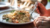 Close-up of a person eating a plate of fried rice with vegetables using a knife and fork, with natural sunlight streaming in and a blurred background of a dining setting.”